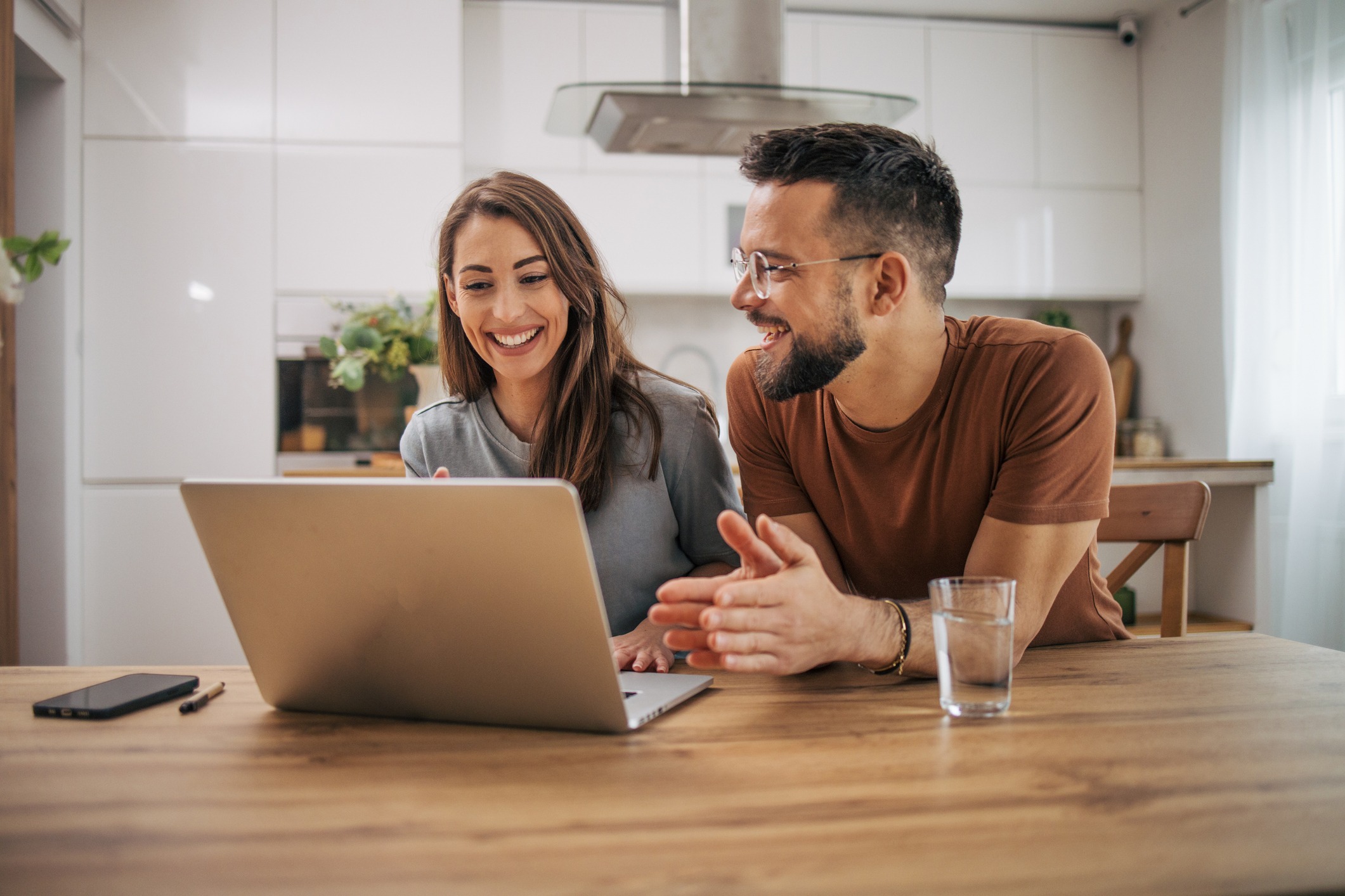 A man and woman sit at a wooden table, smiling and looking at a laptop screen. The man gestures with his hands while the woman types. A glass of water and a smartphone are nearby. The background shows a modern kitchen with white cabinets and a range hood.