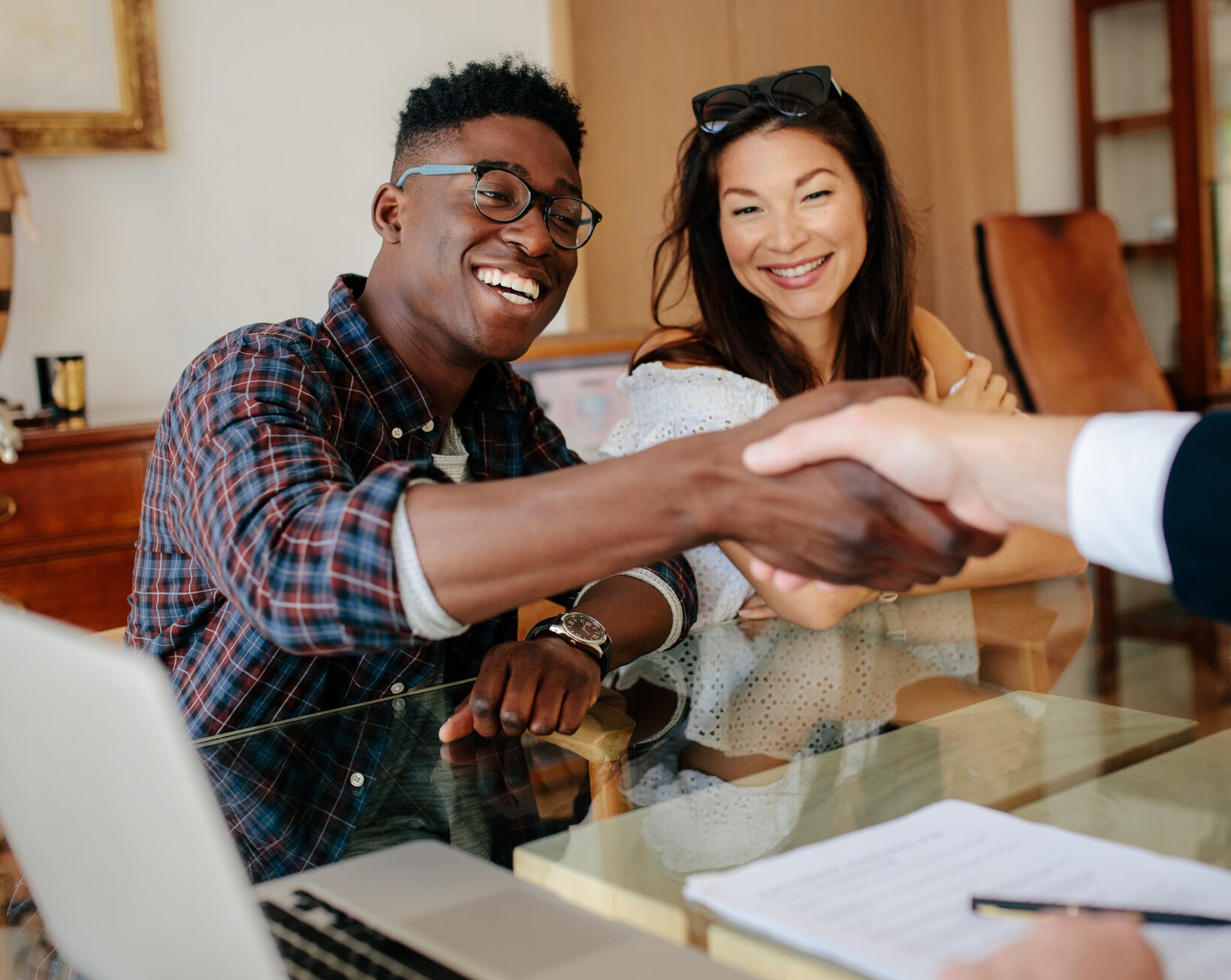 A smiling couple sits at a glass table with a laptop and paperwork. The man, wearing glasses and a plaid shirt, is shaking hands with an unseen person, while the woman, wearing sunglasses on her head and a white blouse, looks on smiling.