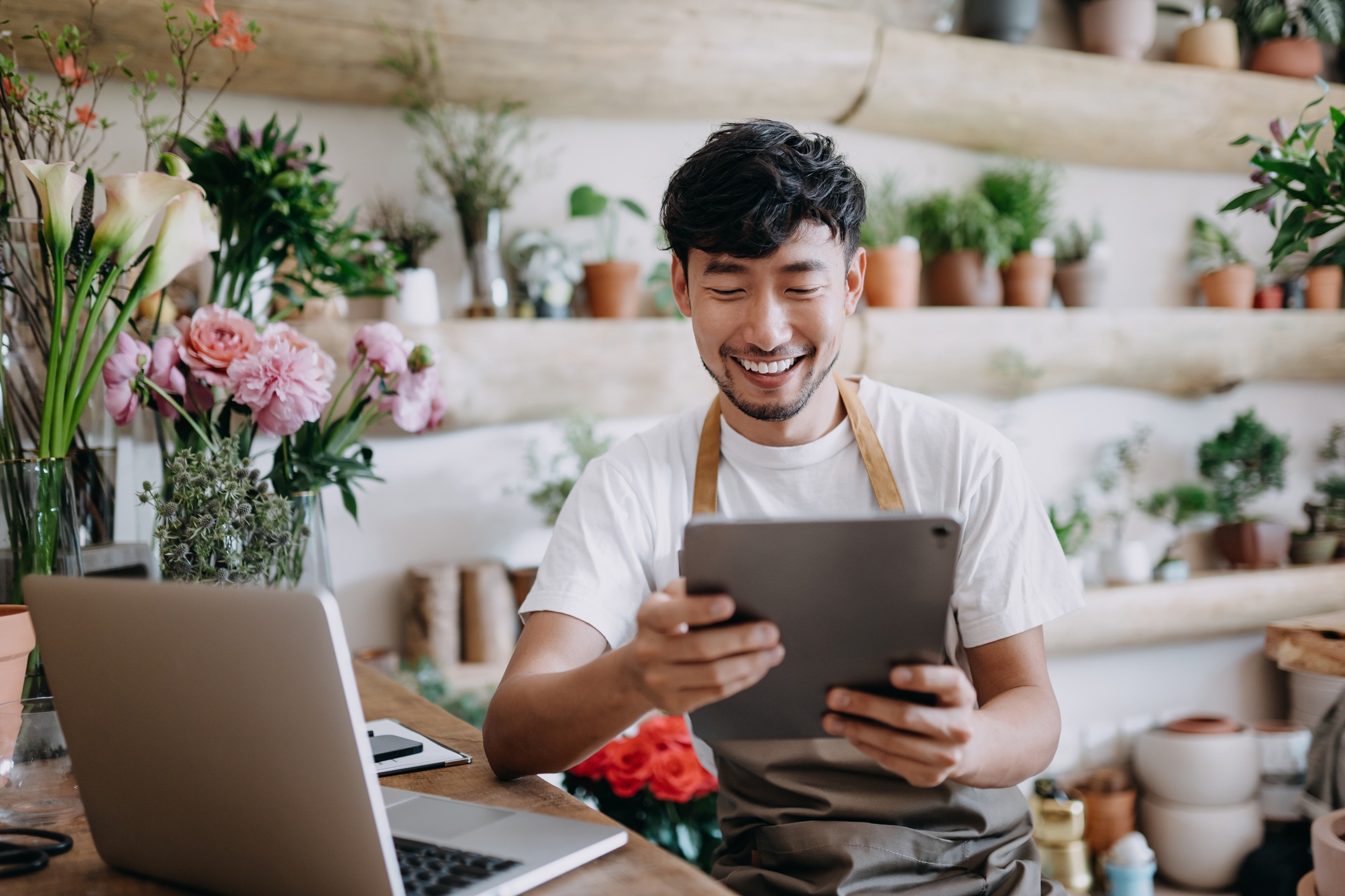 A smiling florist wearing an apron is sitting at a wooden table surrounded by flowers and plants. He is holding a tablet and has a laptop open in front of him, with shelves of potted plants in the background.