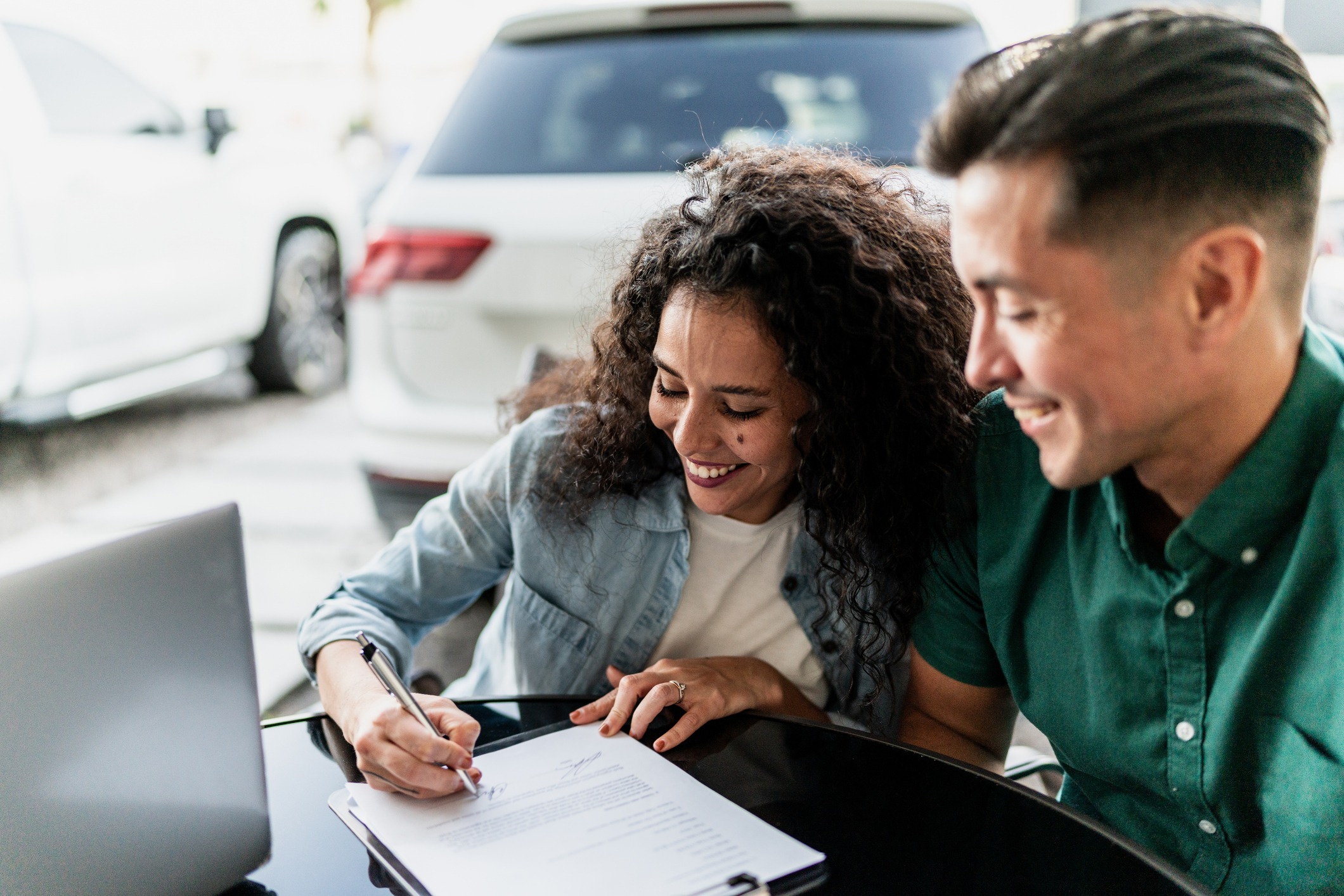 A smiling couple sits at an outdoor table with a laptop and a contract. The woman signs the document while the man looks on. Cars are visible in the background.