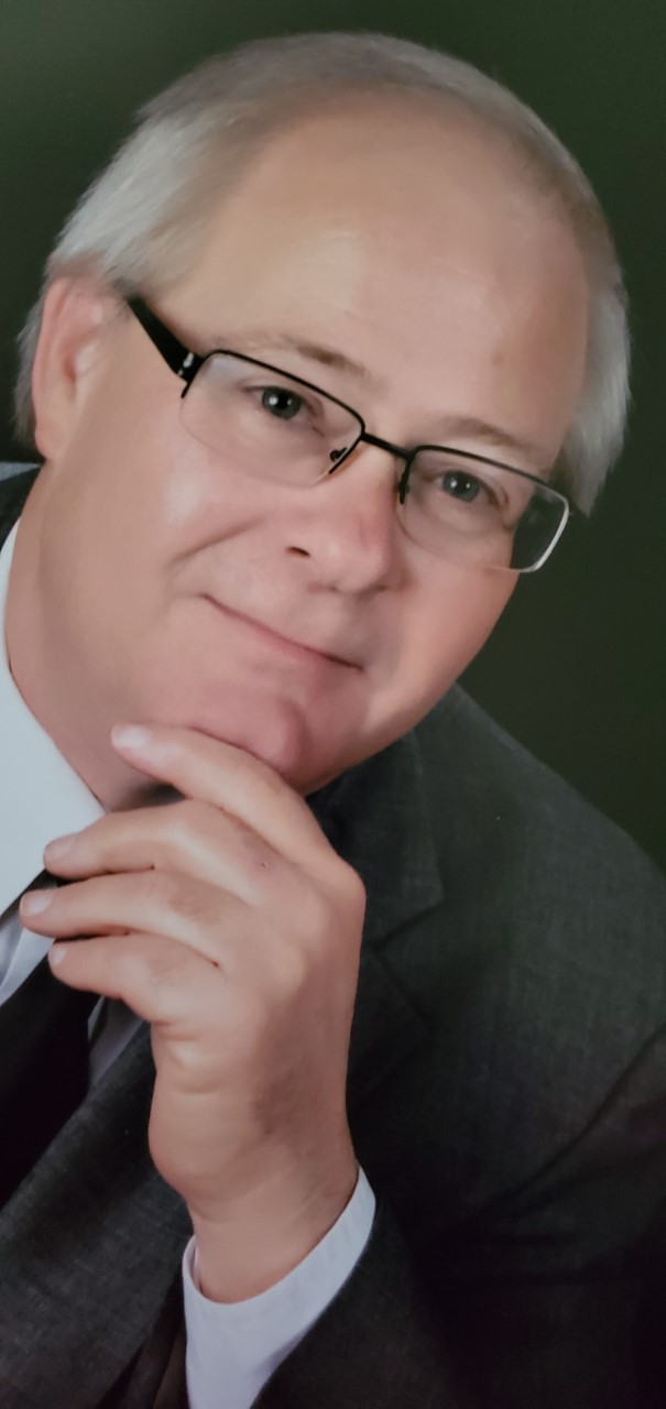 A man with gray hair and glasses wearing a gray suit and tie poses with his chin resting on his hand, looking thoughtfully at the camera against a dark background.