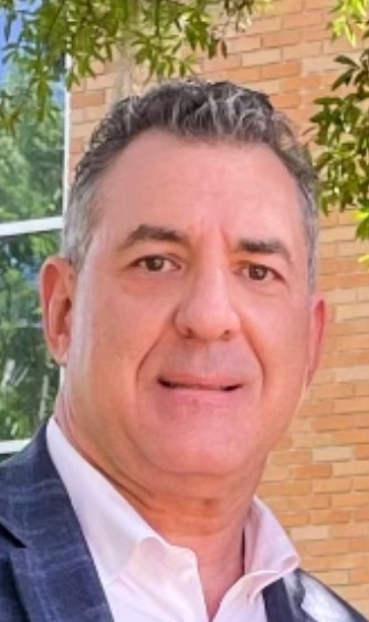 A man with short dark hair, wearing a white shirt and checkered blazer, is standing outdoors. The background features a brick wall, a window reflecting trees, and some greenery.