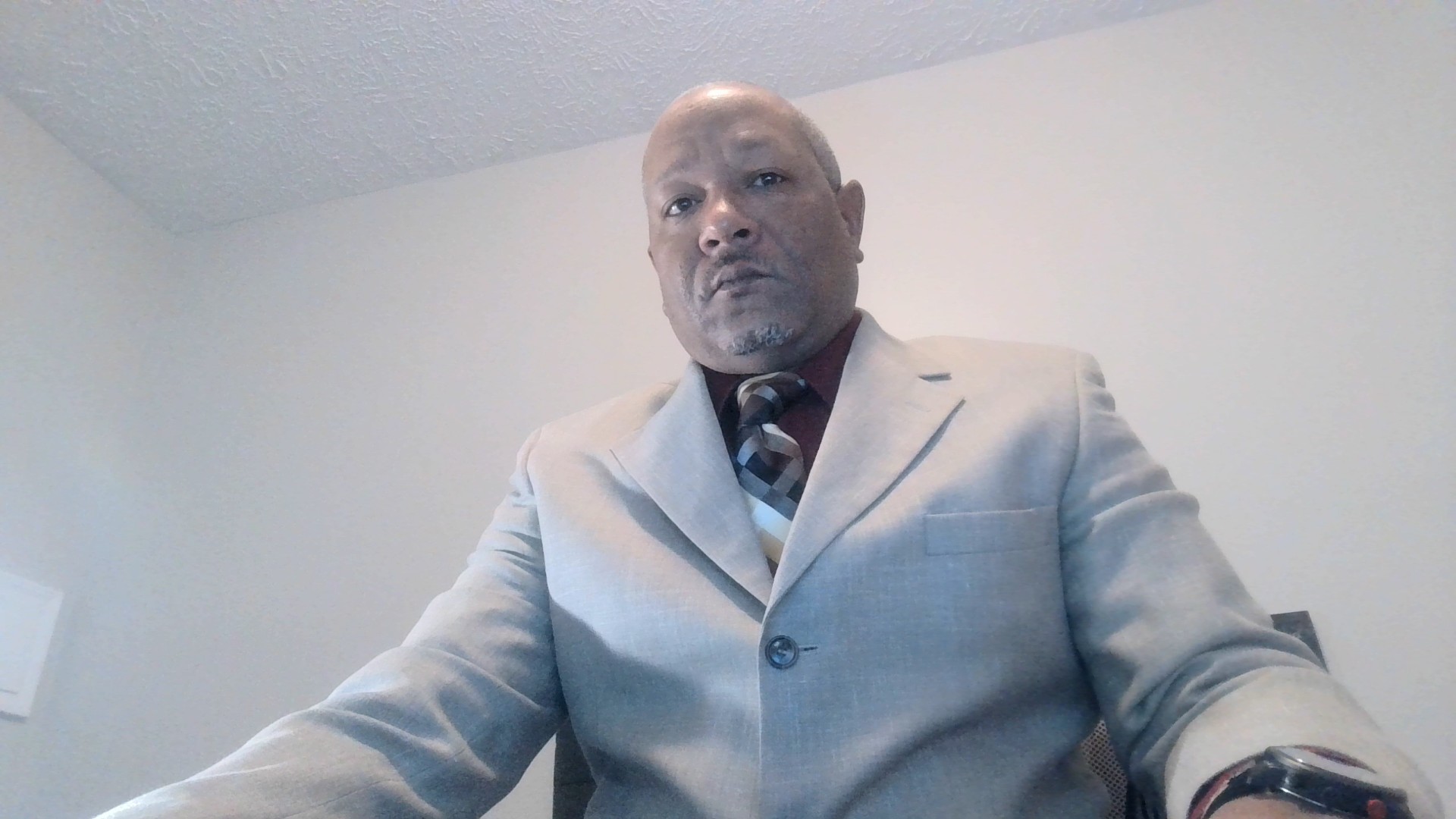 A man in a beige suit and checkered tie sits in an office. The background is a plain, light-colored wall. He looks directly at the camera with a neutral expression.