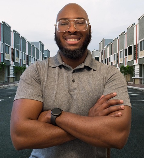 A man with a beard, wearing glasses and a gray polo, smiles with arms crossed. He stands on a street lined with modern townhouses.