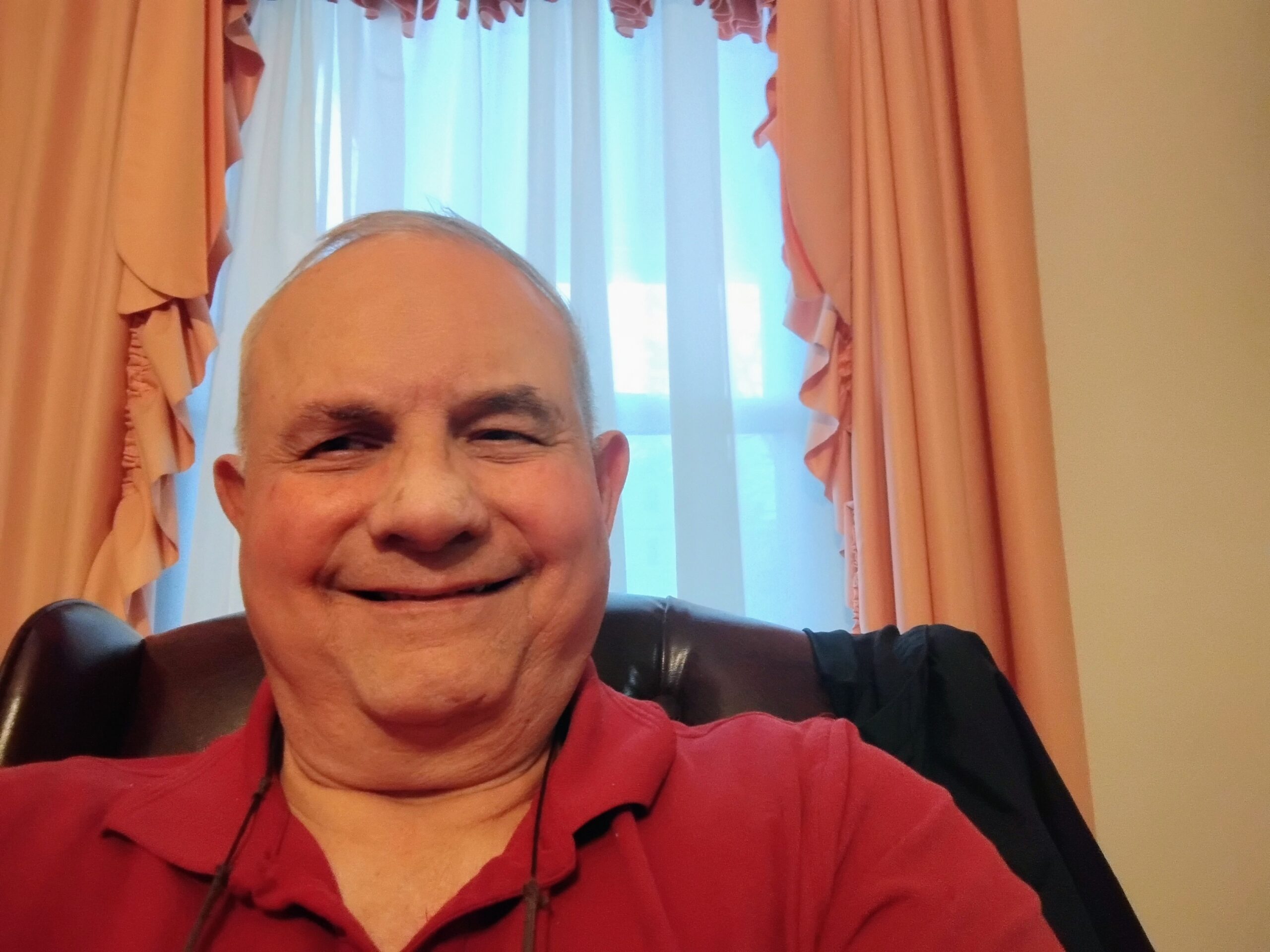 A man wearing a red polo shirt is smiling while sitting in front of a window with sheer white curtains and peach-colored drapes.