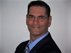 A person with short dark hair and glasses, wearing a dark suit and light blue shirt, smiling at the camera against a plain white background.