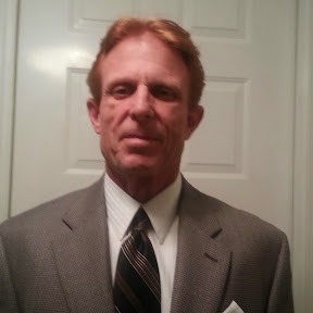 A middle-aged man with short reddish hair wearing a gray suit, white dress shirt, and striped tie, standing in front of a white door and looking at the camera.