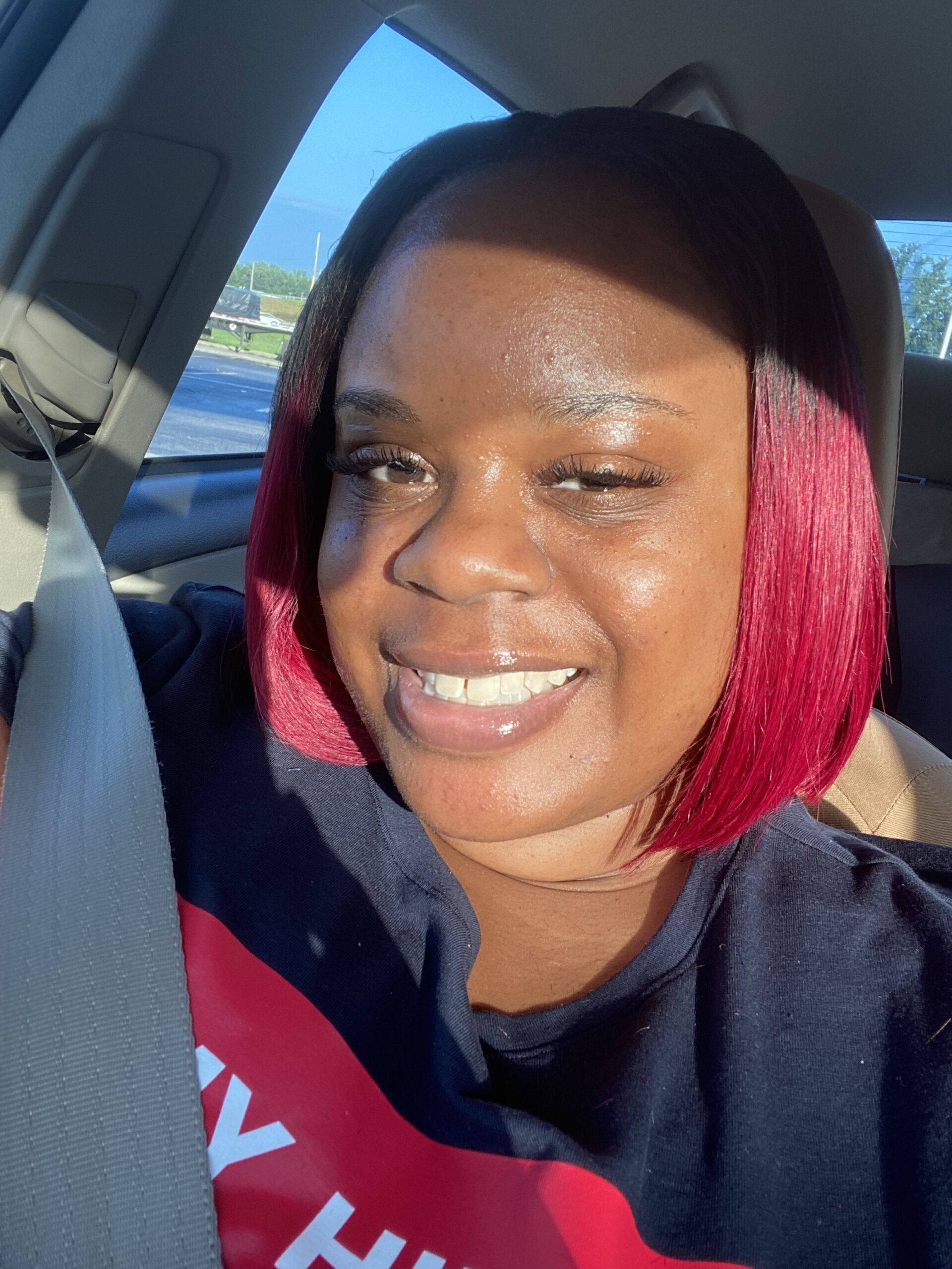 A woman with straight, shoulder-length red hair smiles while sitting in a car. Sunlight shines on her face, and she is wearing a black shirt with a seatbelt across her chest.