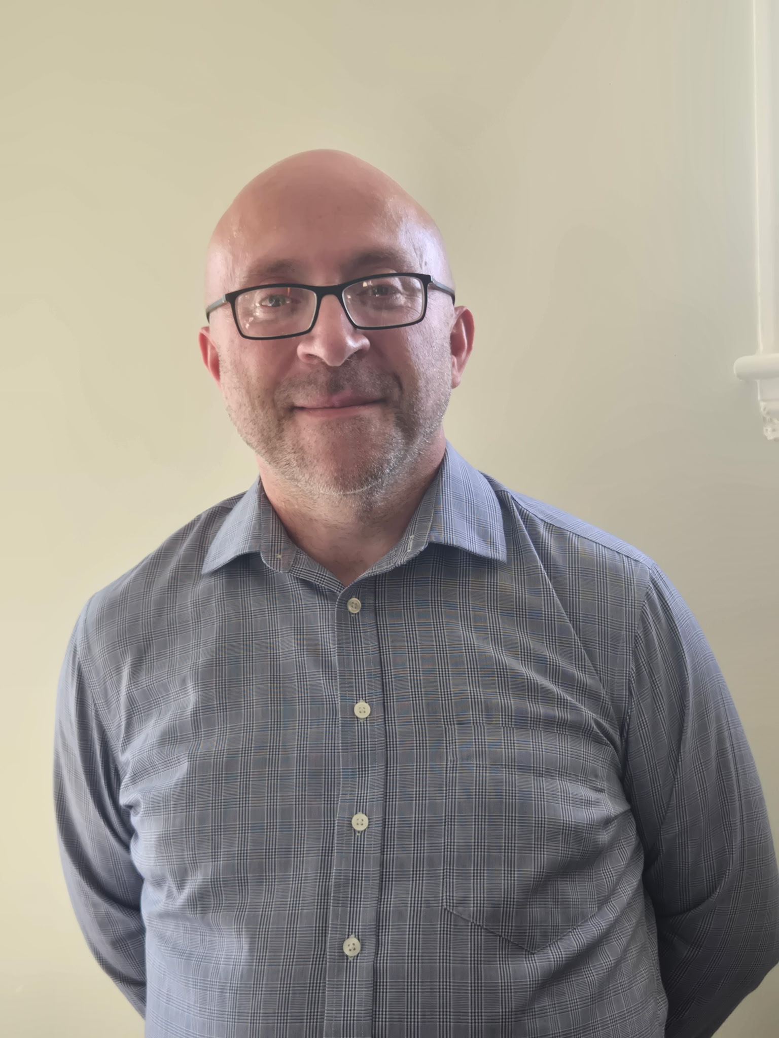 A middle-aged man with glasses, a bald head, and a short beard is smiling slightly. He is wearing a blue, checkered button-up shirt and standing against a plain, light-colored wall.