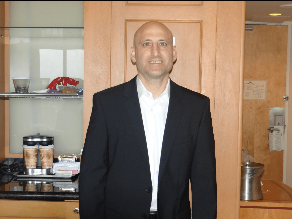 A man in a black suit and white shirt stands indoors near a wooden cabinet and countertop with snacks and coffee supplies. A hotel room door and ice bucket are visible in the background.