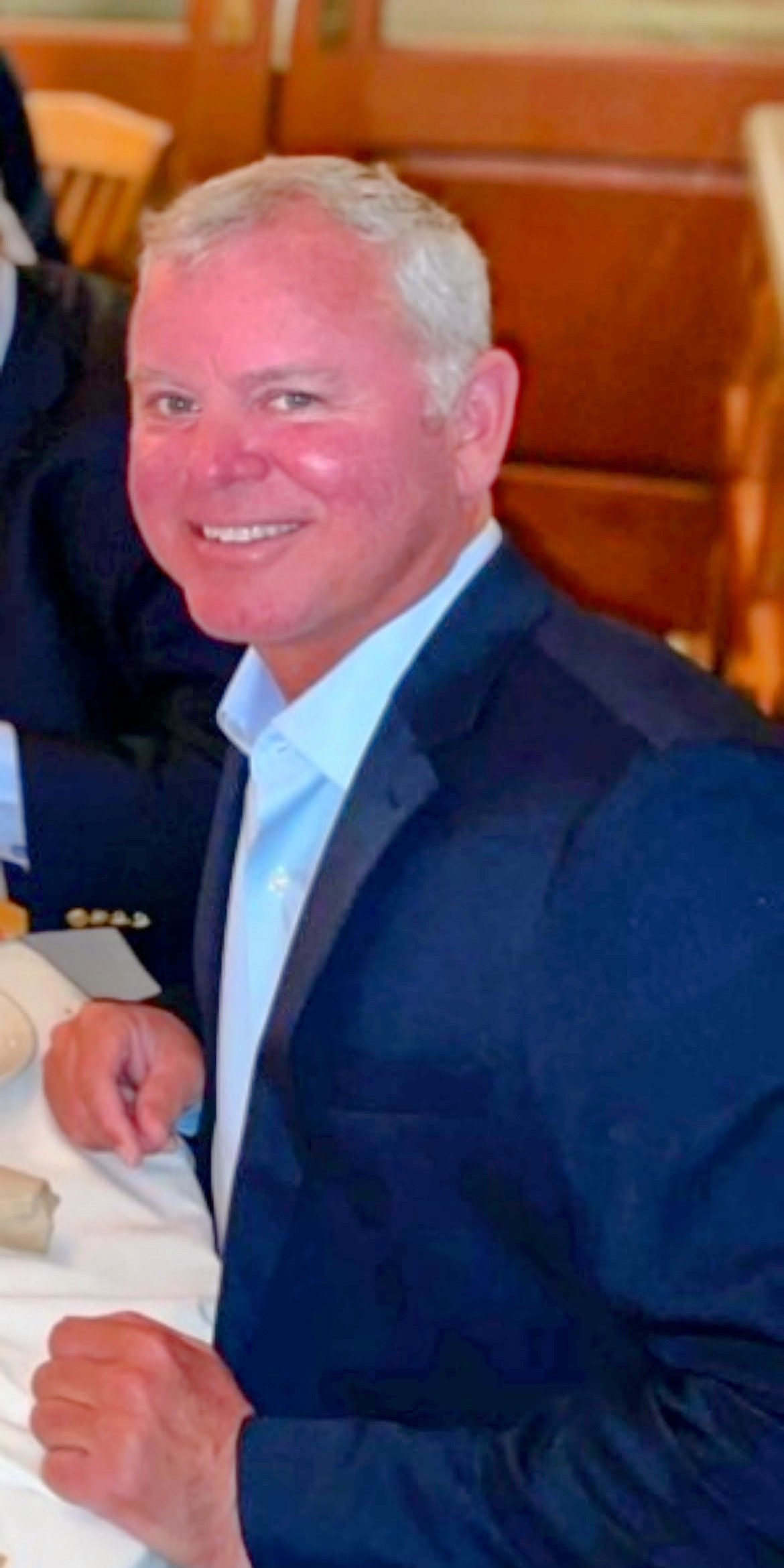 A man with short gray hair, wearing a dark suit jacket and light blue shirt, is smiling while seated at a table in a restaurant setting.