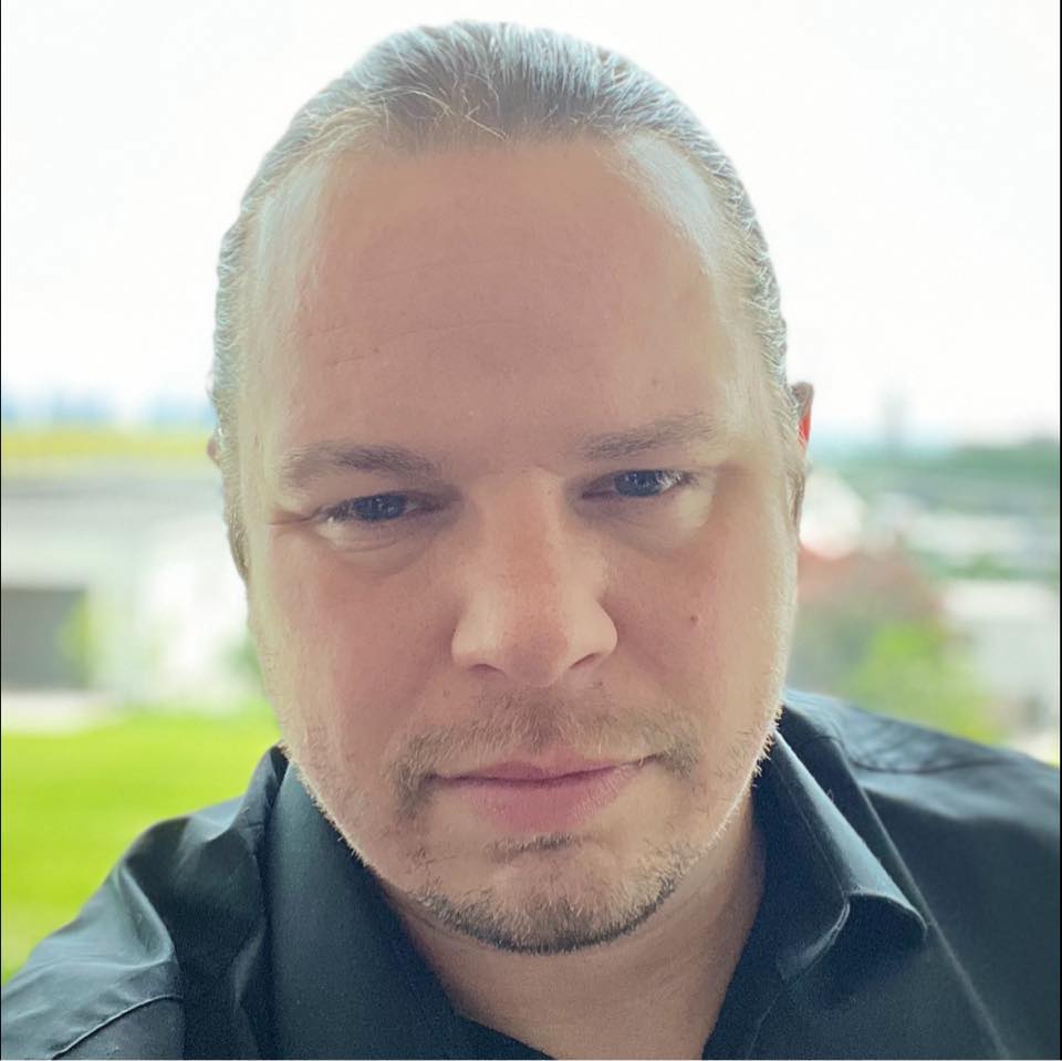 A person with fair skin and light brown hair tied back is wearing a black shirt and looking at the camera. The background is bright with greenery and blurred outdoor scenery.