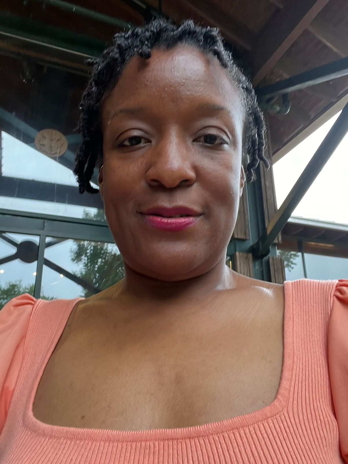 A person with short, twisted hair and wearing a peach-colored top is smiling slightly at the camera. They are outdoors near a building with glass windows and a wooden roof.