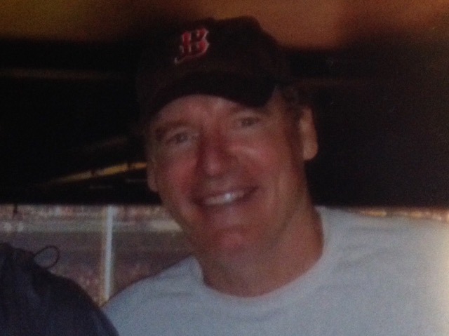 A man wearing a black baseball cap with a red "B" logo and a light-colored shirt smiles at the camera. The background is dimly lit and out of focus.