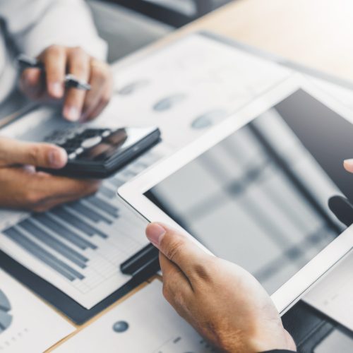 Two people are working together at a desk. One person is holding a smartphone and the other is pointing at a tablet. They have financial documents with charts and graphs spread out on the desk in front of them.